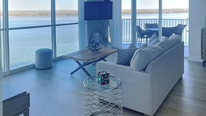 Un salon spacieux doté d’une grande fenêtre donnant sur un plan d’eau, d’un canapé blanc, d’une petite table et d’un téléviseur fixé au mur.