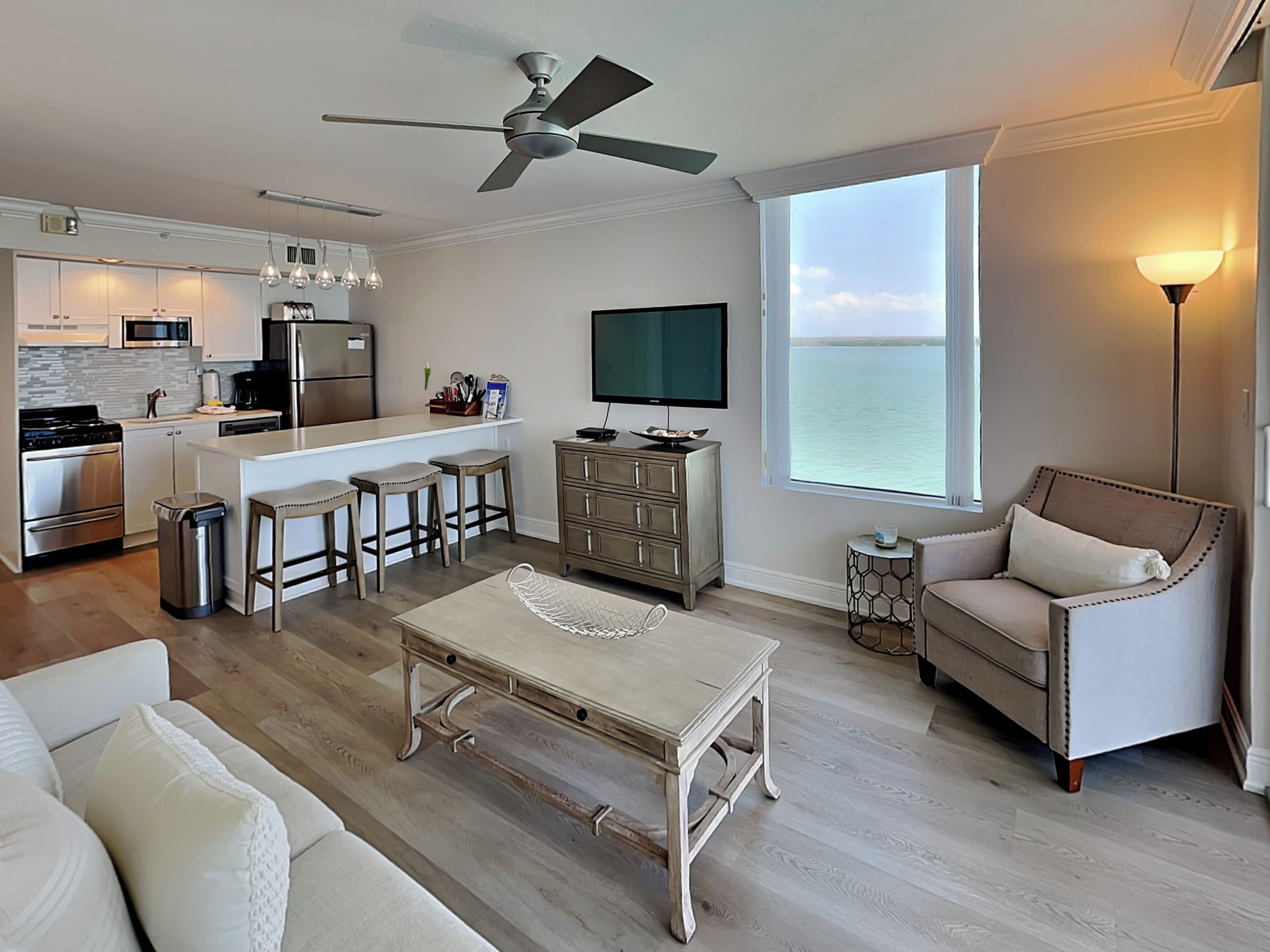 A modern kitchen with a central island, bar stools, a dining table, and a sofa. A television is mounted on the wall, and there is a large window with a view of the sea.