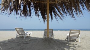 Una playa con tres sillas de playa blancas bajo un paraguas de techo de paja, orientada al océano.
