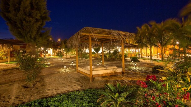 Una escena nocturna de un resort con un pabellón de techo de paja, palmeras y un jardín bien iluminado.