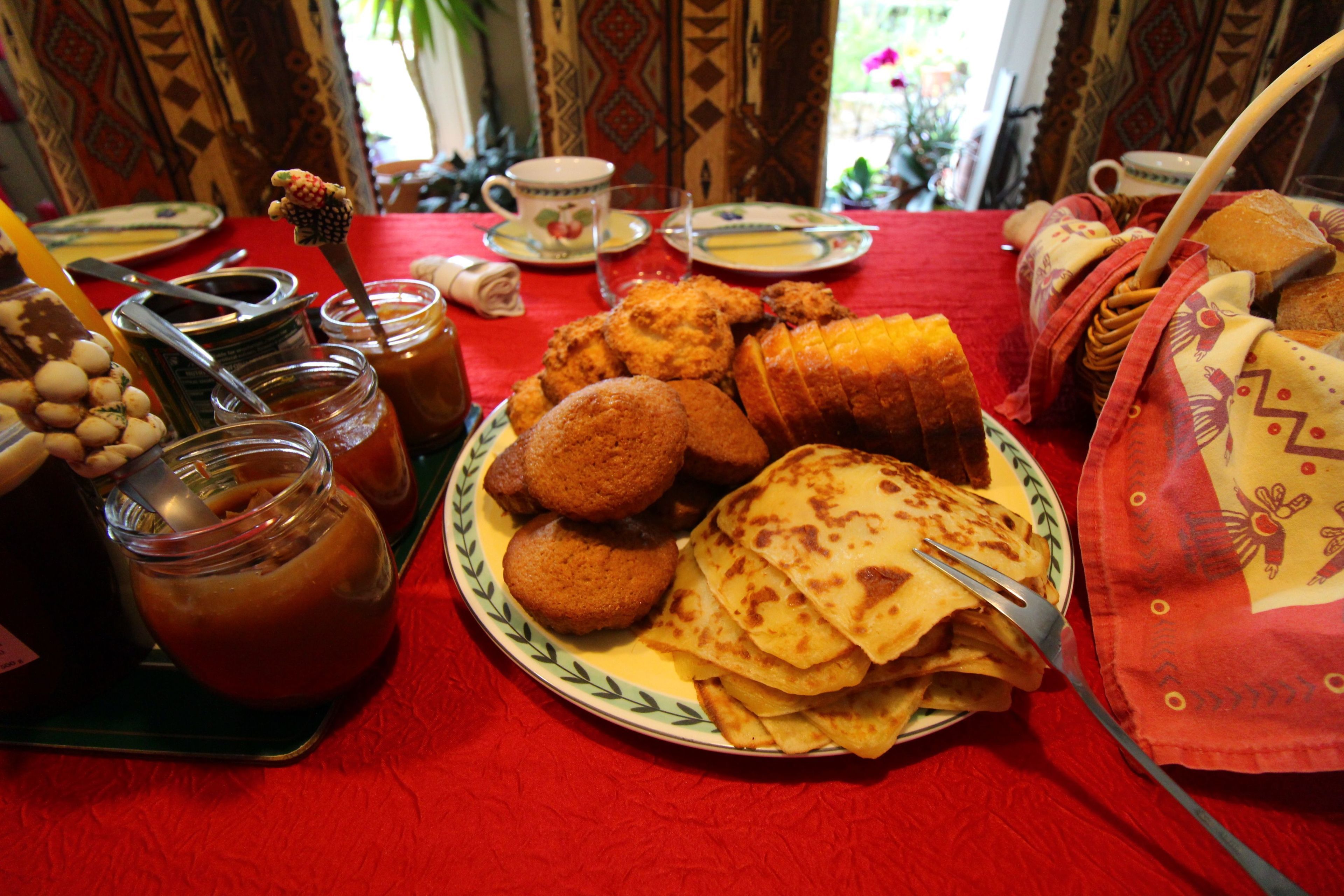 Une table recouverte d’une nappe rouge, une assiette remplie de nourriture, notamment des crêpes, un pot de confiture et une tasse.