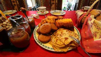 Une table recouverte d’une nappe rouge, une assiette remplie de nourriture, notamment des crêpes, un pot de confiture et une tasse.