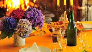 A table set for dining with a vase of flowers, candles, and a bottle of wine, with a fireplace in the background. - Ti Er Mad (Kernascleden)