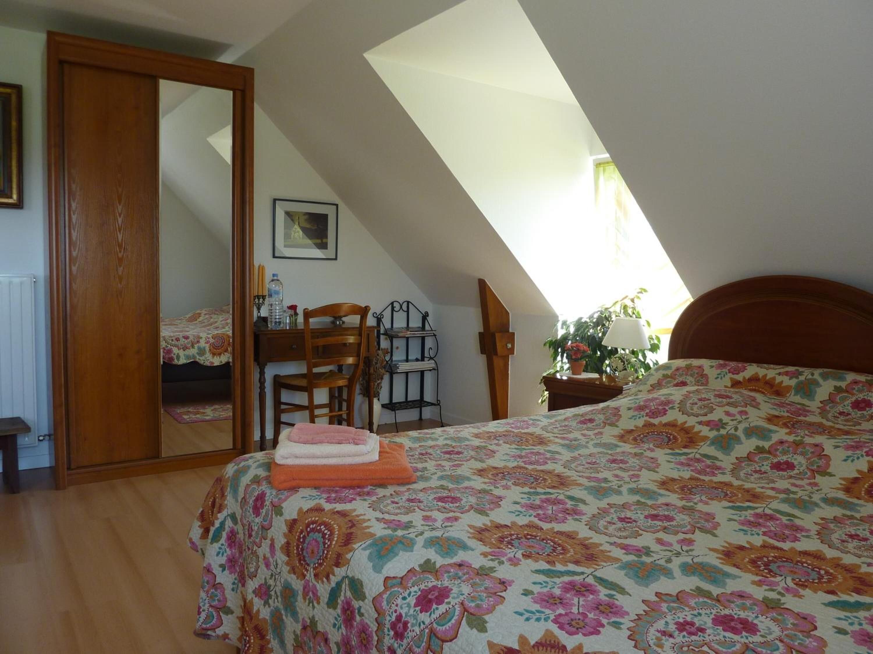 Quarto com teto inclinado, cama com colcha floral, escrivaninha com cadeira, espelho e janela que proporciona luz natural.