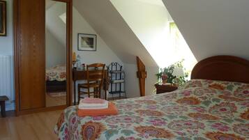Quarto com teto inclinado, cama com colcha floral, escrivaninha com cadeira, espelho e janela que proporciona luz natural.