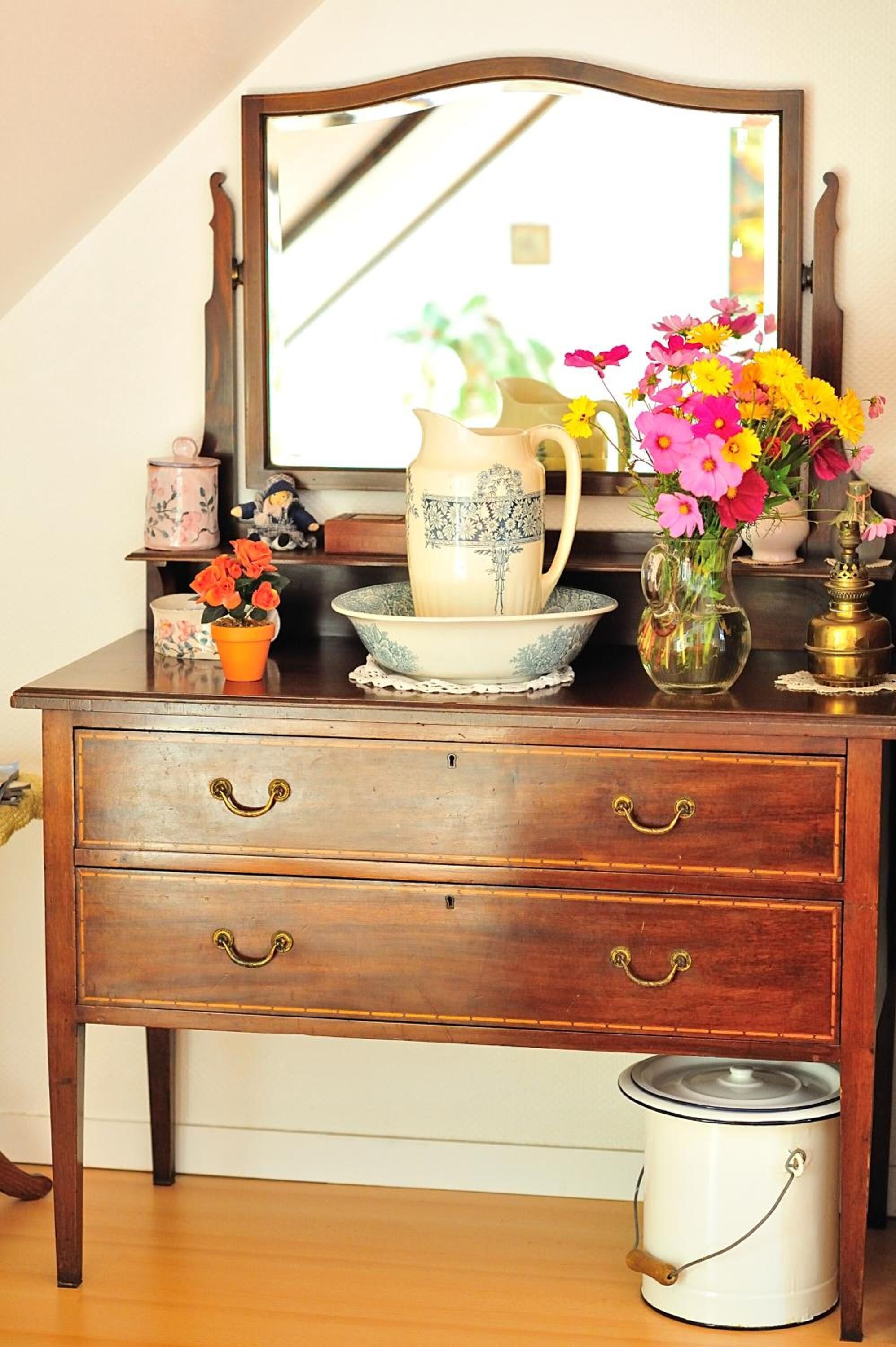 Een houten dressoir met drie lades, een spiegel, en diverse decoratieve objecten, waaronder een vaas met bloemen, een theepot en een klein beeldje.