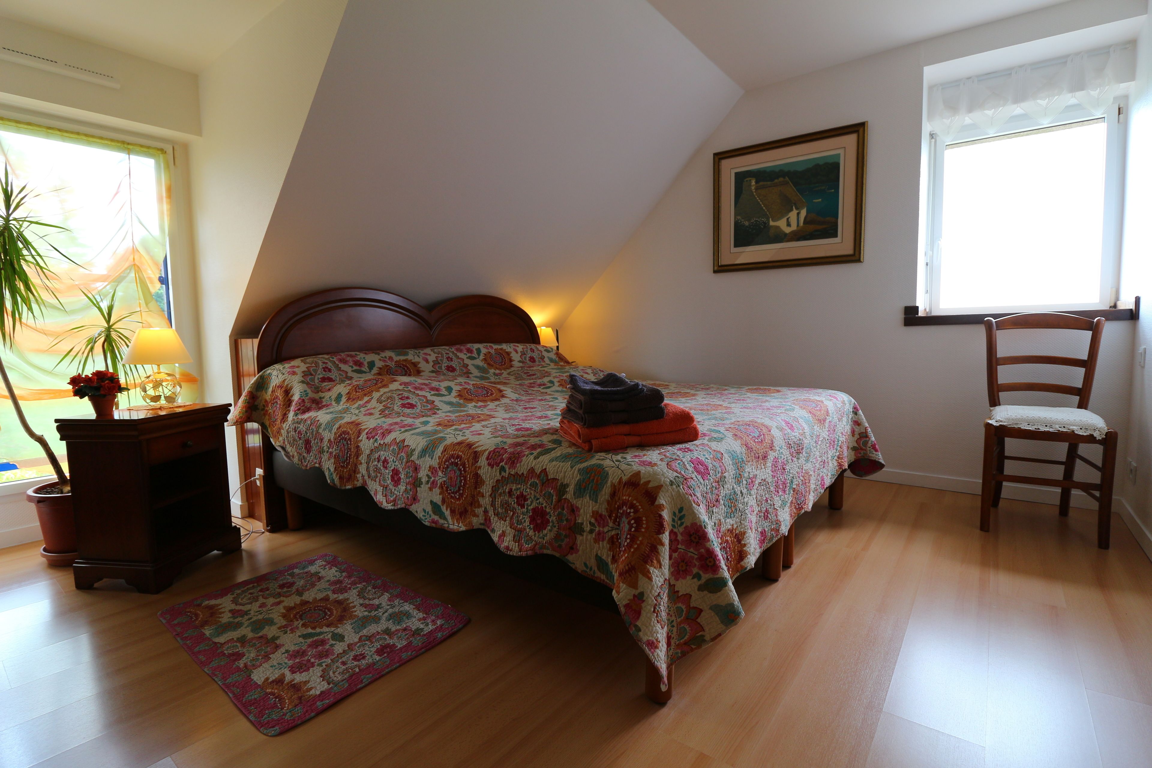 A bedroom with a bed, a chair, a nightstand, a lamp, a window, and a painting on the wall.