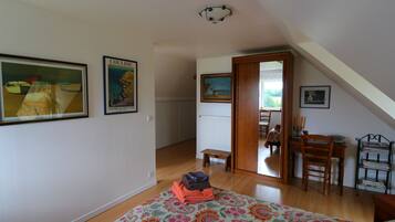 A room with a wooden floor, a bed with a floral bedspread, a small bench, a dining table with chairs, and a bookshelf.