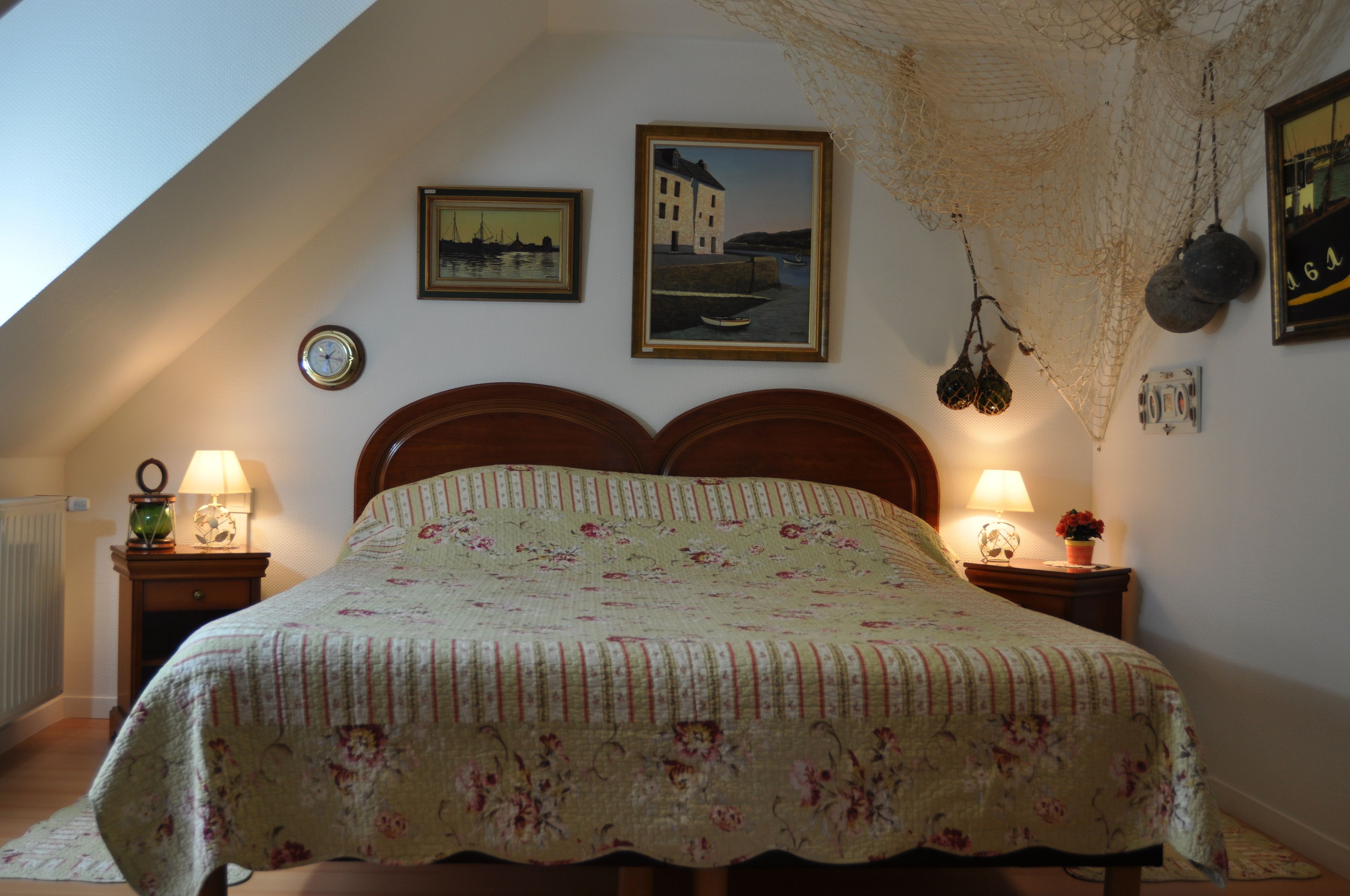 A bedroom with a bed, two bedside tables, a wall clock, two framed pictures, and a hanging net.