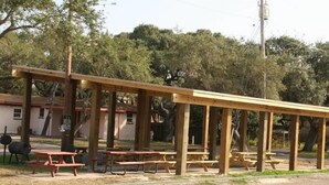 A covered picnic area with wooden benches and picnic tables, surrounded by trees and a building in the background. - Bayfront Cottages (Rockport)