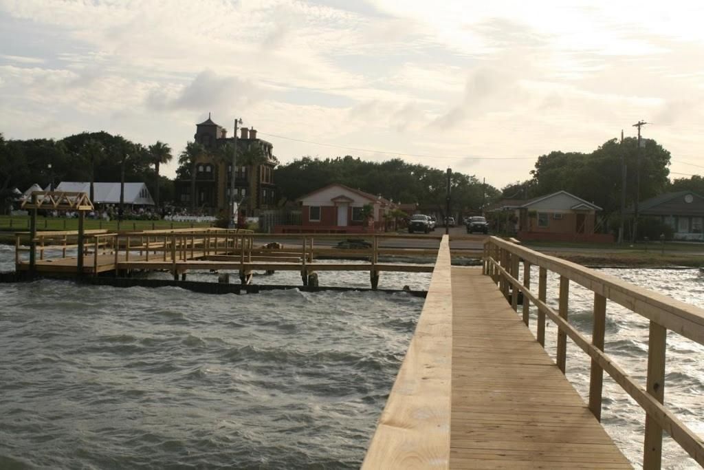 Un muelle de madera que se adentra en una masa de agua, con edificios y árboles al fondo.