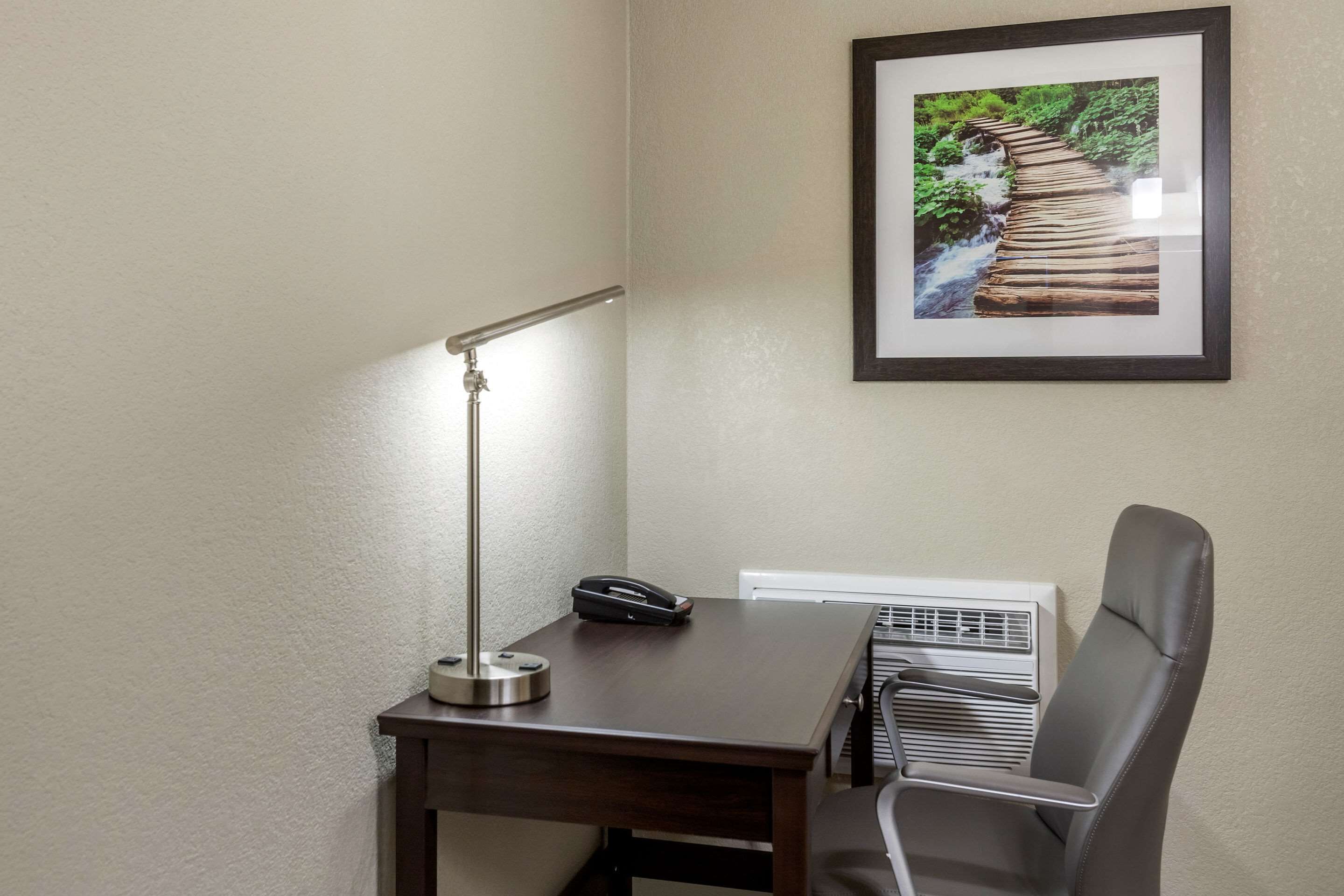 Escritório no canto com uma mesa, uma cadeira, luminária de mesa, quadro emoldurado e unidade de ar condicionado.