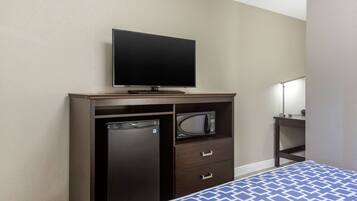 A hotel room with a bed, a TV mounted on a wooden cabinet, a microwave, and a small table with a lamp.