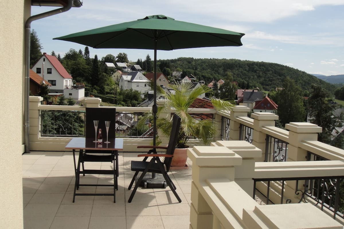 A balcony with a table and chairs, a green umbrella, and a view of houses and hills in the background.