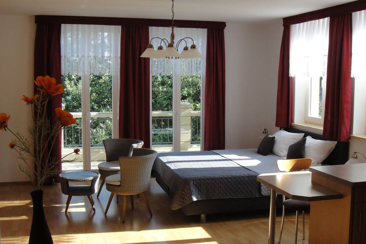 A bedroom with a bed, bedside table, chair, and a small desk. There is a window with curtains, a vase with flowers, and a hanging light fixture.