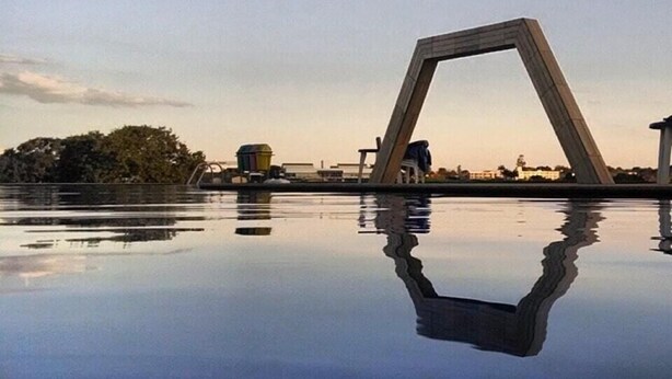 Un puente con una estructura geométrica singular reflejado en aguas tranquilas, rodeado de árboles y edificios al fondo.