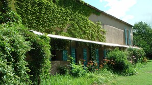 A rustic building with a covered veranda, surrounded by lush greenery and flowering plants. - Cottage with private terrace and view of the park! (Apt)