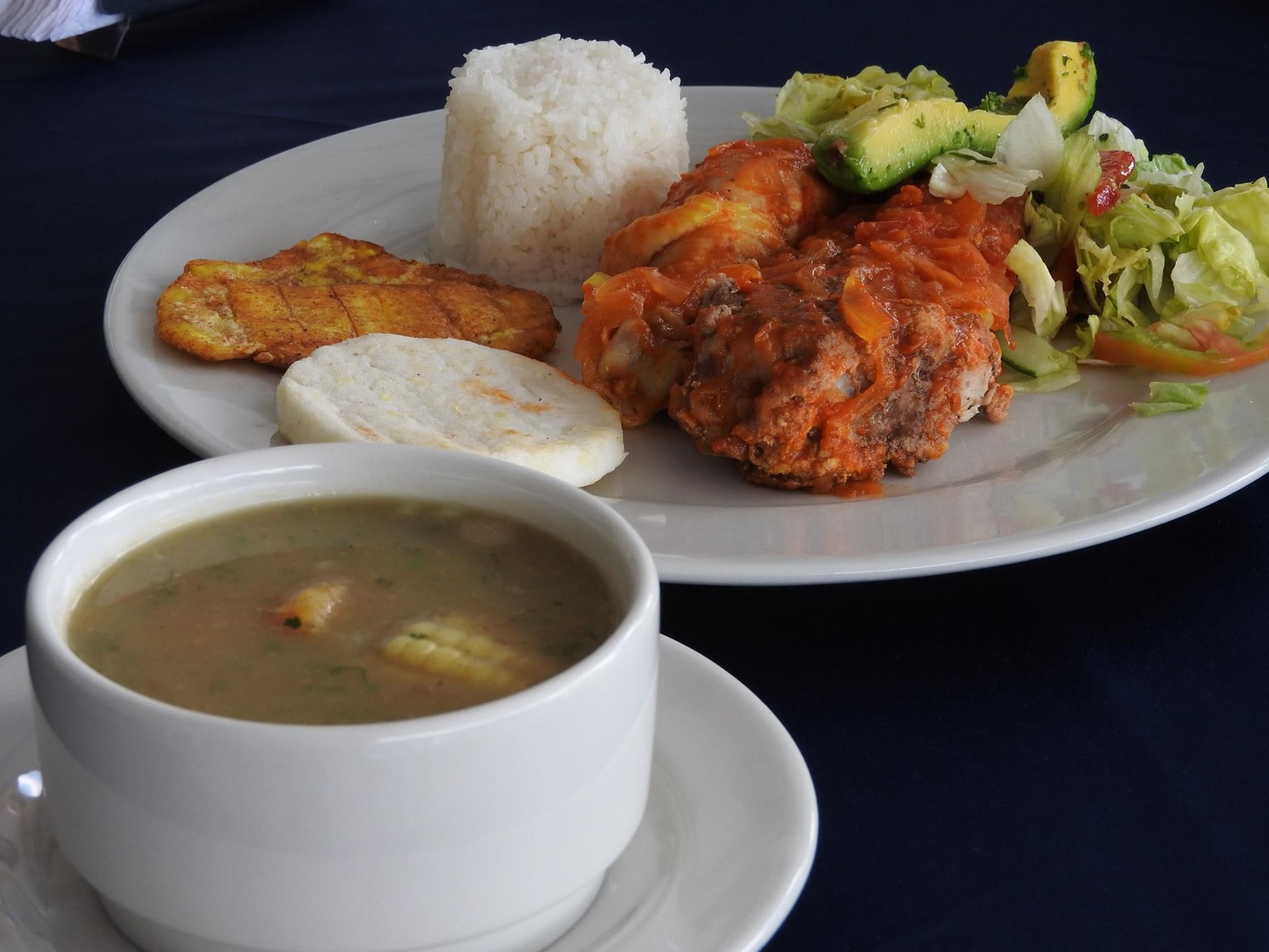 A plate with rice, fried plantains, a meat dish, avocado salad, and a bowl of soup.