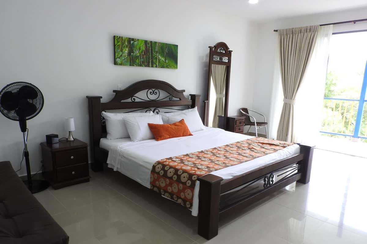 A neatly arranged bedroom with a large bed, a bedside table, a standing fan, and a window with curtains.