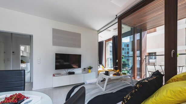 Una habitación de hotel moderna con cama, televisión, zona de comedor y balcón con vista a la ciudad.