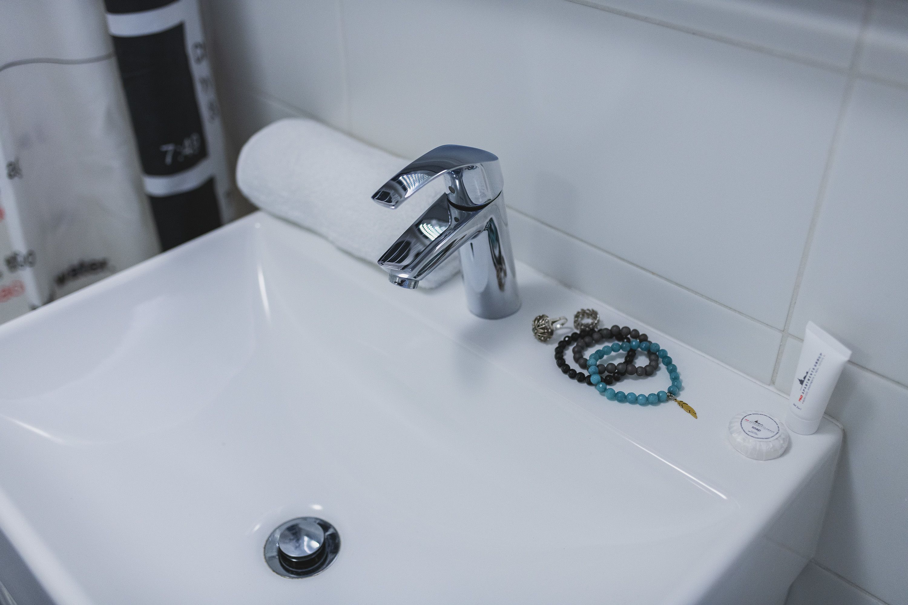 Ein Badezimmerwaschbecken mit chromfarbenem Wasserhahn, Abfluss und persönlichen Gegenständen wie Armbändern und einer Creme-Tube.