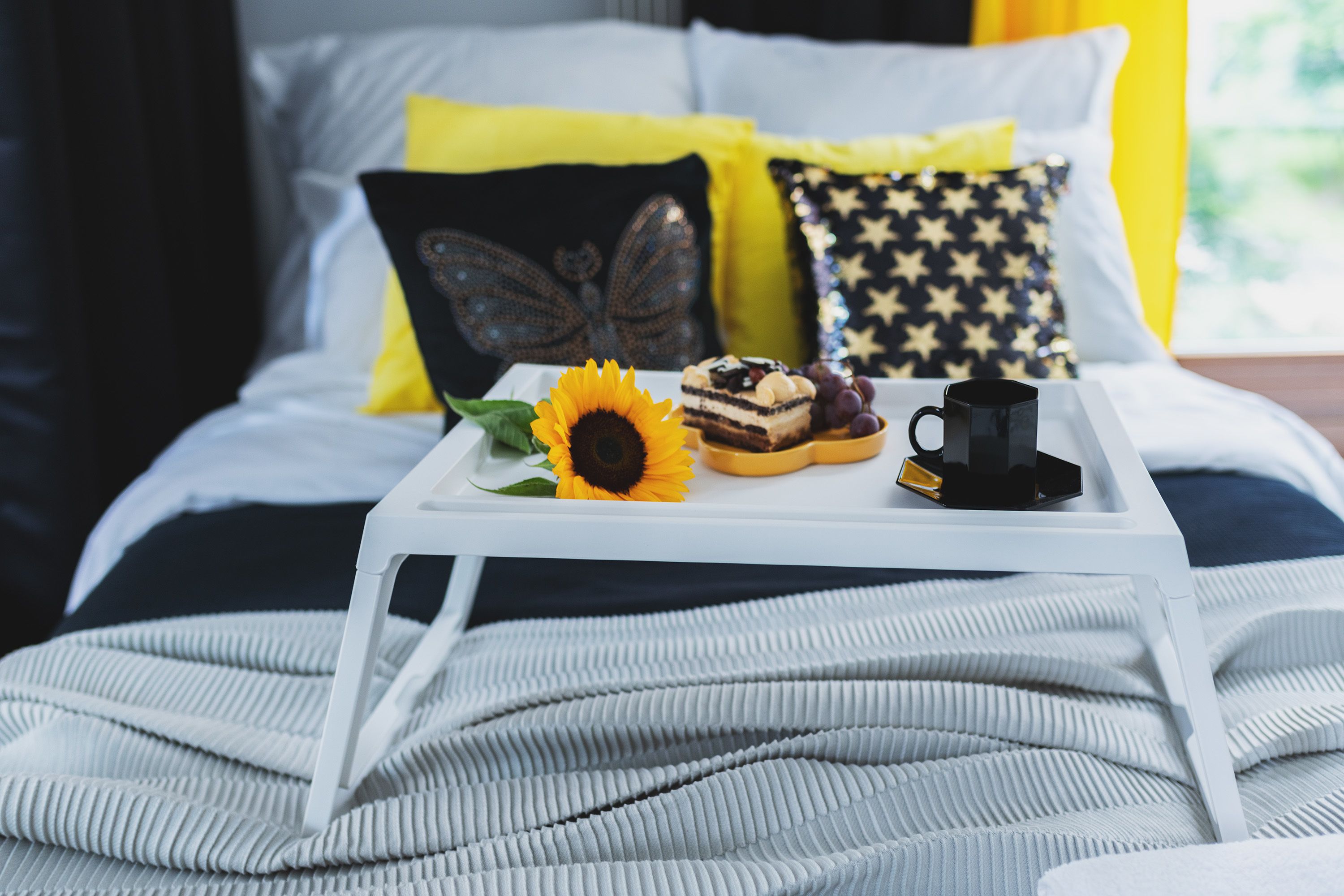 A bed with a white tray holding a sunflower, a slice of cake, grapes, and a black mug.