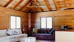 A cozy cabin interior with wooden walls, a sofa, a couch, ottomans, a guitar, and a kitchenette.