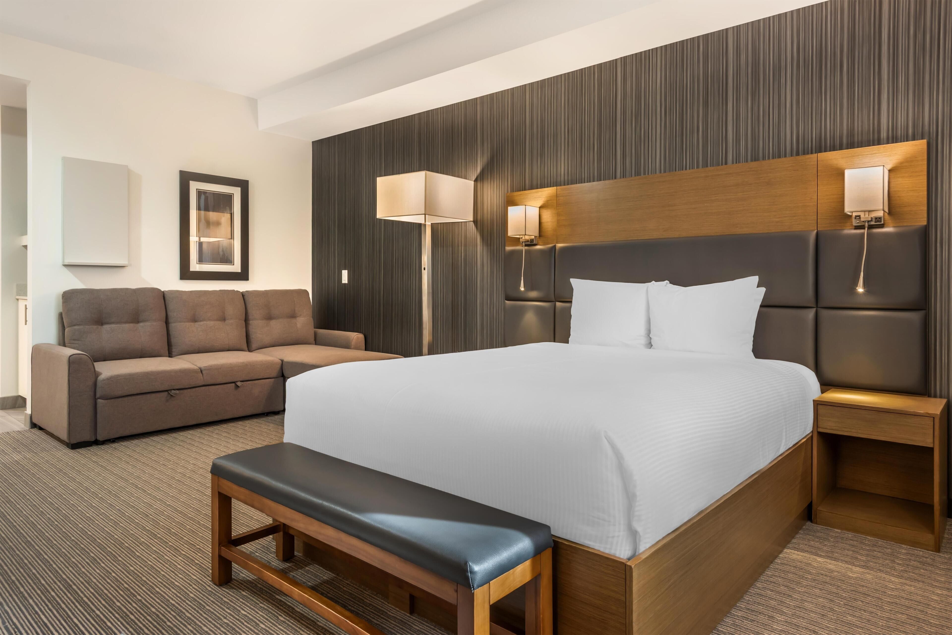 Una habitación de hotel con una cama grande, un banco, un sofá, dos lámparas fijadas a la pared y un cuadro enmarcado.