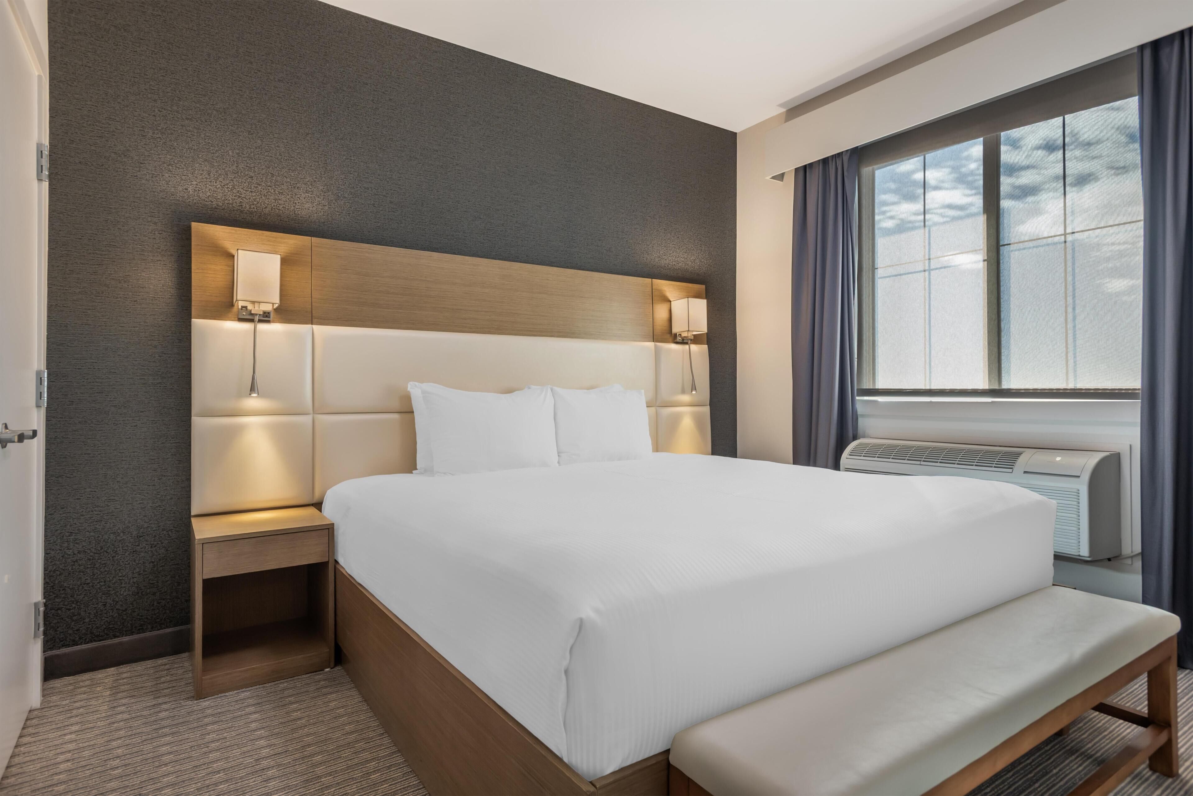 Una habitación de hotel con una cama grande, lámparas de noche, un banco, un aire acondicionado de pared y una ventana con cortinas.