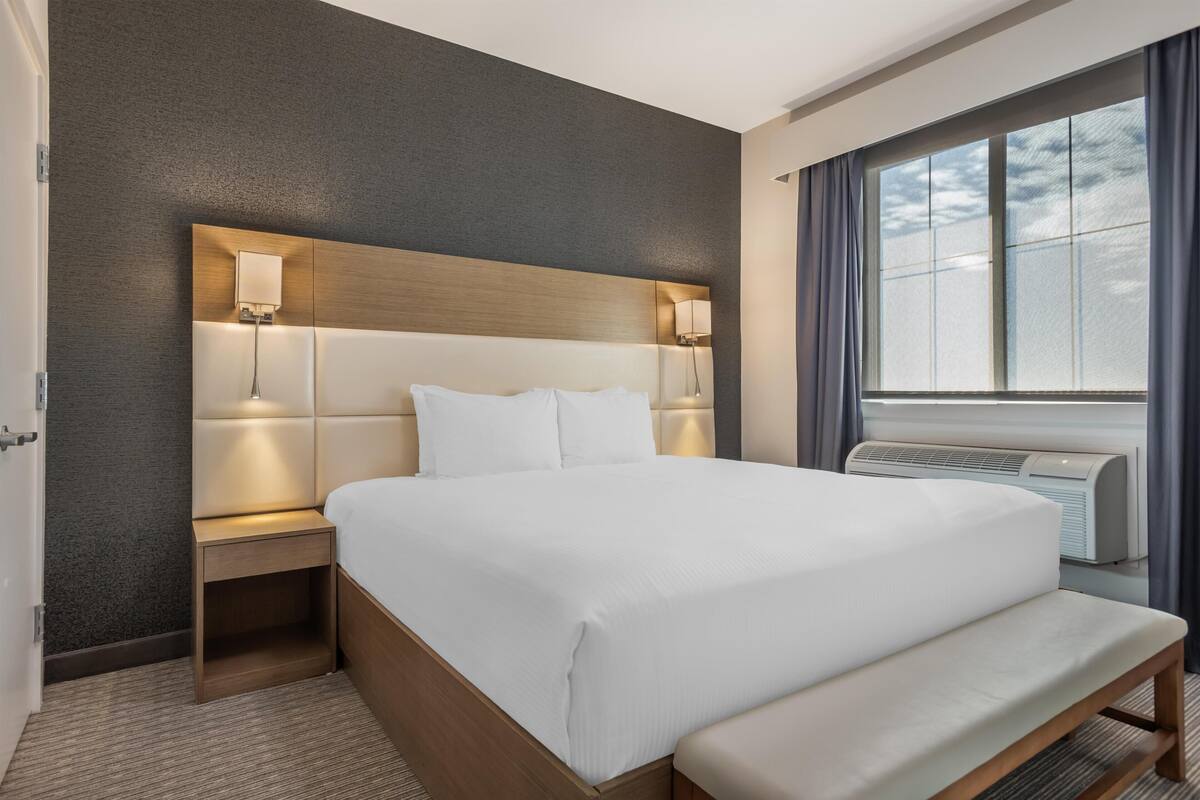Una habitación de hotel con una cama grande, lámparas de noche, un banco, un aire acondicionado de pared y una ventana con cortinas.