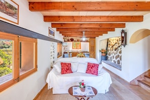 Un dormitorio con cama, una mesita con un florero, vigas de madera, una ventana y una escalera.