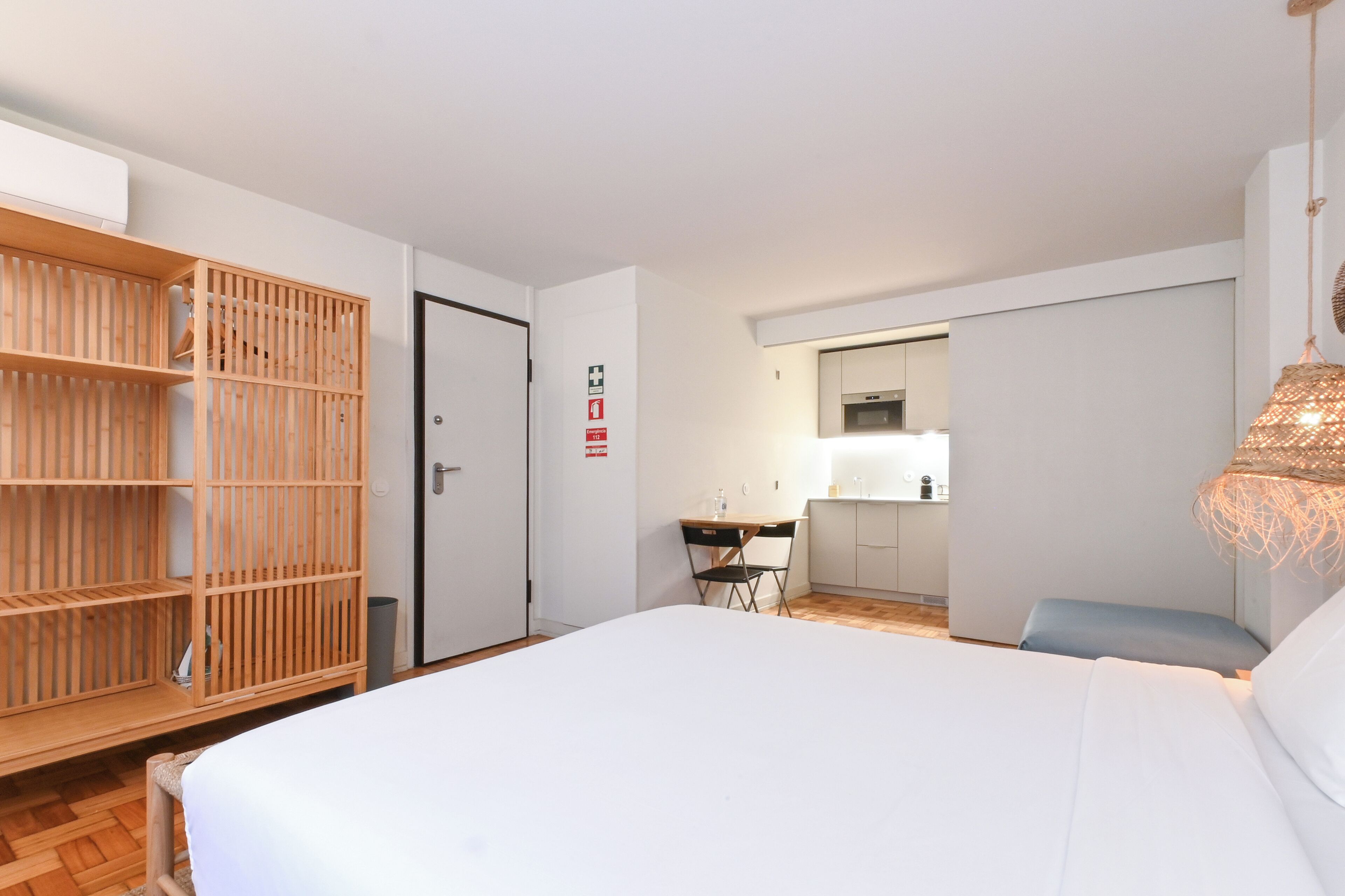 A hotel room with a bed, a wooden wardrobe, a desk with a chair, and a kitchenette in the background.