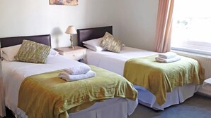 Een hotelkamer met twee eenpersoonsbedden, elk met een witte dekbedovertrek en een groene plaid. Tussen de bedden staat een nachtkastje met een lamp, en aan de rechterkant een raam met gordijnen.