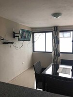 Una habitación con una mesa de comedor y sillas, un televisor montado y una ventana con cortinas.