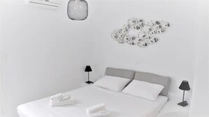 A minimalist bedroom with a large bed, two bedside tables with lamps, a wall-mounted light fixture, and a decorative wall piece. - Traditional Cycladic Home with a large private pool with sea view (Mikonos)