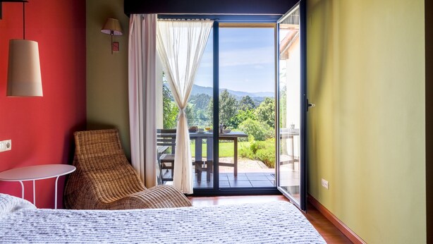A bedroom with a bed, a wicker chair, a small table, and a view of a garden through a sliding glass door. - Apto. 3. La mar salada (Castiello De La Marina)