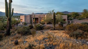 A desert-style building with a flat roof, surrounded by cacti and dry grass, with mountains in the background. - Southwest home w/ Hot Tub, heated* pool, and amazing Mtn. Views! (Cave Creek)