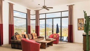 A spacious living room with a sofa, armchair, and coffee table, large windows with curtains, a ceiling fan, and a cactus plant. - Southwest home w/ Hot Tub, heated* pool, and amazing Mtn. Views! (Cave Creek)