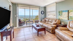 A living room with a large TV, leather sofas, a coffee table, and a view of the ocean through a sliding glass door. - Beach & Tennis 2005 (Bonita Springs)