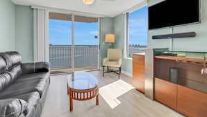 A modern hotel room with a balcony, a leather sofa, a glass coffee table, a wooden cabinet, and a flat-screen TV.