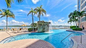 A swimming pool with a waterfall feature, surrounded by lounge chairs and palm trees, with a view of the water and a building in the background.