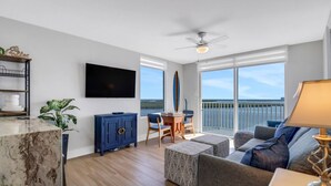 A modern living room with a flat-screen TV, a ceiling fan, a dining area with a table and chairs, and a large window with a view of the water. - Lovers Key Resort 1003 (Fort Myers Beach)