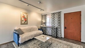 A living room with a blue sofa, a white furry throw, a glass coffee table, patterned rugs, a framed floral artwork, a patterned curtain, a black metal console table, and a wooden door. - Cottage w/ Patio & Grill - 25 Min to Taos Valley! (El Prado)