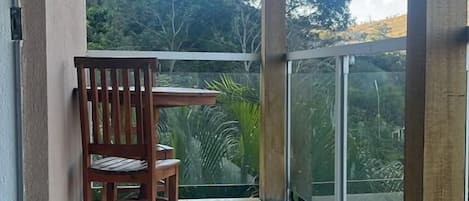 A covered balcony with a wooden table and chairs, a glass door leading to a lush green landscape.
