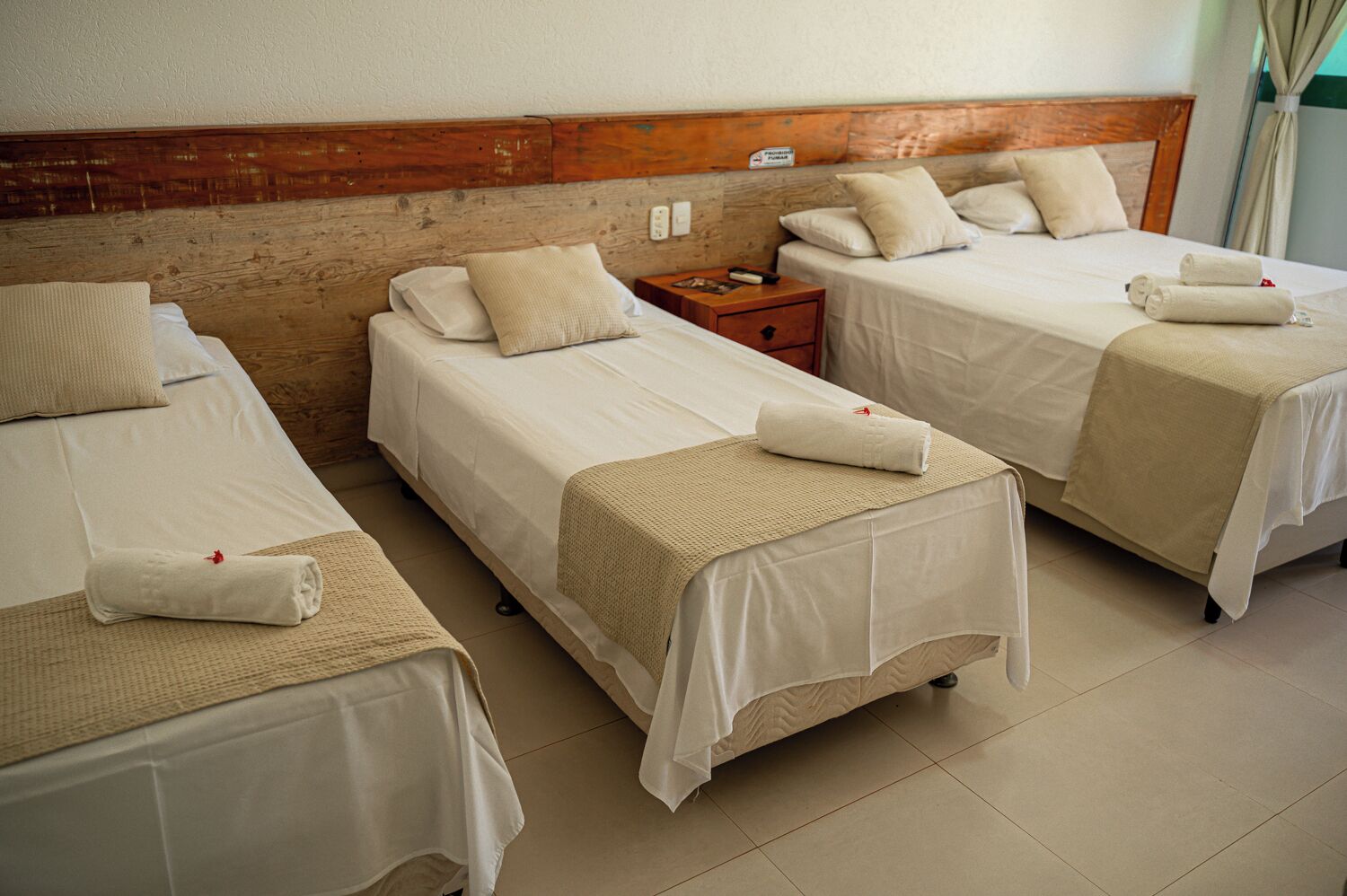 Habitación de hotel con tres camas individuales, cada una con una colcha blanca y una manta color beige. Se observa un cabecero de madera, una mesita de noche con lámpara y un cartel en la pared.