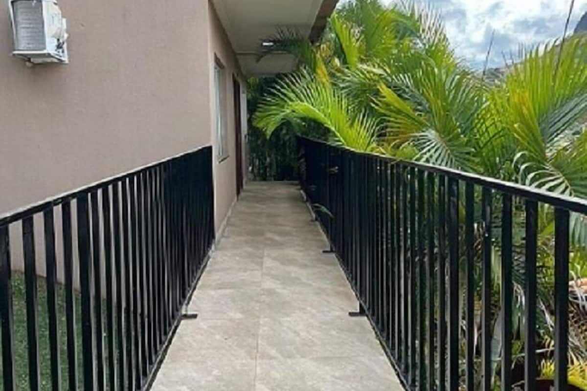 Un pasillo con suelo de baldosas, barandilla de metal negro y vistas a un edificio con balcón y unidad de aire acondicionado.