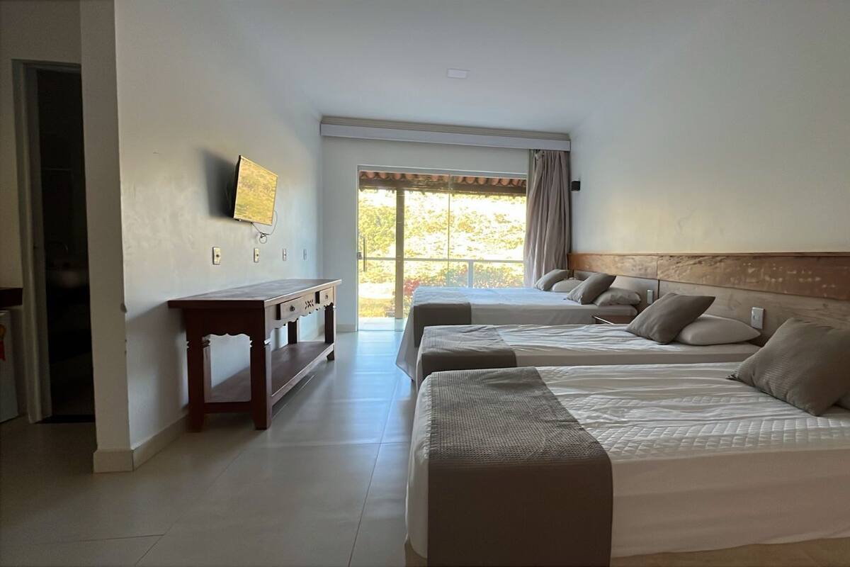 Una habitación de hotel con tres camas, una mesita de noche de madera, un televisor de pantalla plana montado en la pared y un ventanal con cortinas.
