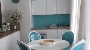 Uma área de refeições com uma mesa redonda e quatro cadeiras, uma cozinha com armários brancos, um relógio e um vaso com flores.