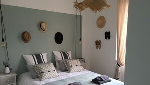 Uma cama bem arrumada com travesseiros e um cobertor, uma mesinha de cabeceira com abajur, e uma parede com chapéus e sapatos.