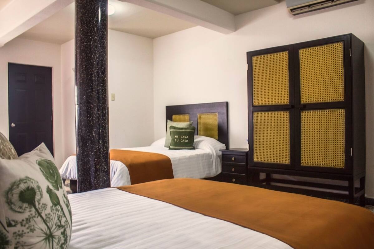 A hotel room with two beds, a large pillar, a wardrobe with a woven front panel, and a door in the background.